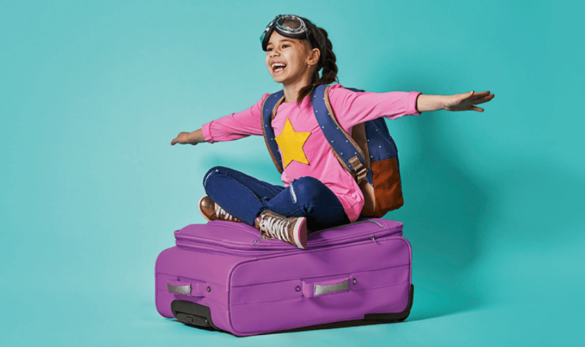 A young girl, full of joy, wearing a pink star shirt and sitting playfully on a purple suitcase, symbolising family travel and fun. 