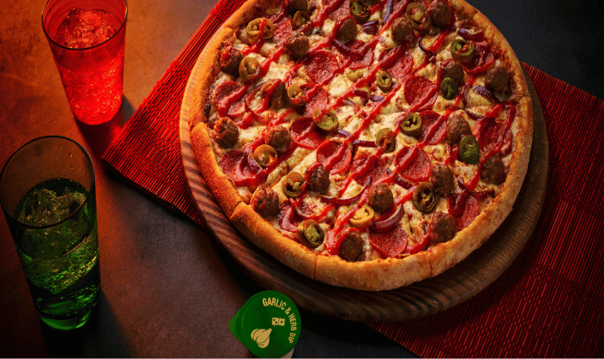 A large Domino's The Ultimate Spicy Meatball pizza sits on top of a wooden board and a red placemat. There are red and green glasses of clear fizzy drinks next to it and a garlic and herb dip pack.