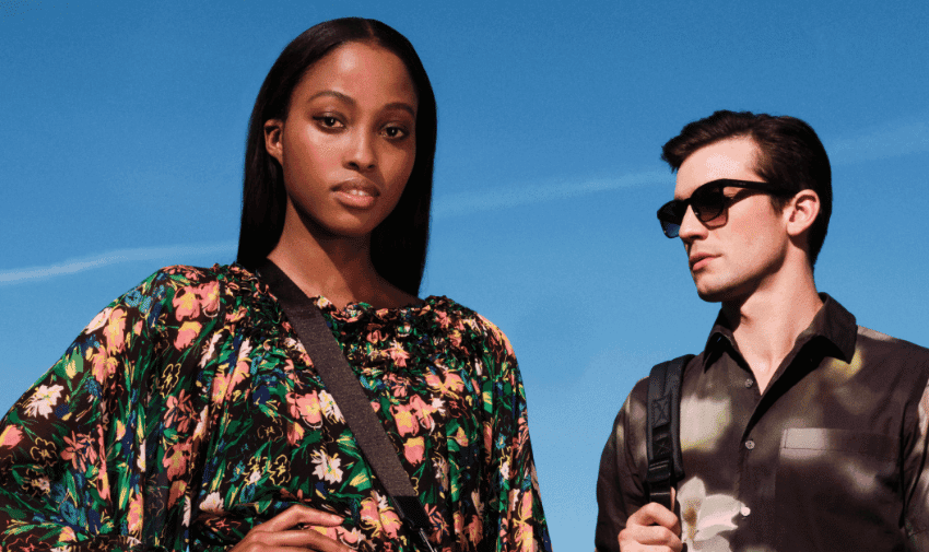 A woman with long straight hair wearing a floral print dress and a crossbody bag stands in the foreground. A man with short dark hair wearing sunglasses and a patterned shirt with a backpack stands slightly behind her. The sky is clear and blue.