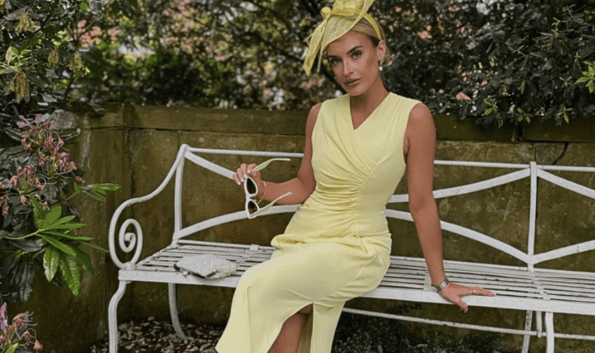 A woman in a sleeveless yellow dress and matching hat sits on a white bench in a garden. She rests one hand on her lap, holding a pair of white sunglasses, and the other on the bench. Greenery and a stone wall form the background.