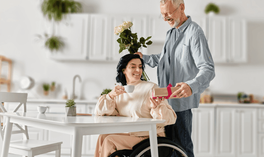 A person in a wheelchair holds a wrapped gift with a red ribbon and bow in their hands. The person wears blue jeans and a light blue shirt. The background is blurred, focusing on the gift and the person's hands.