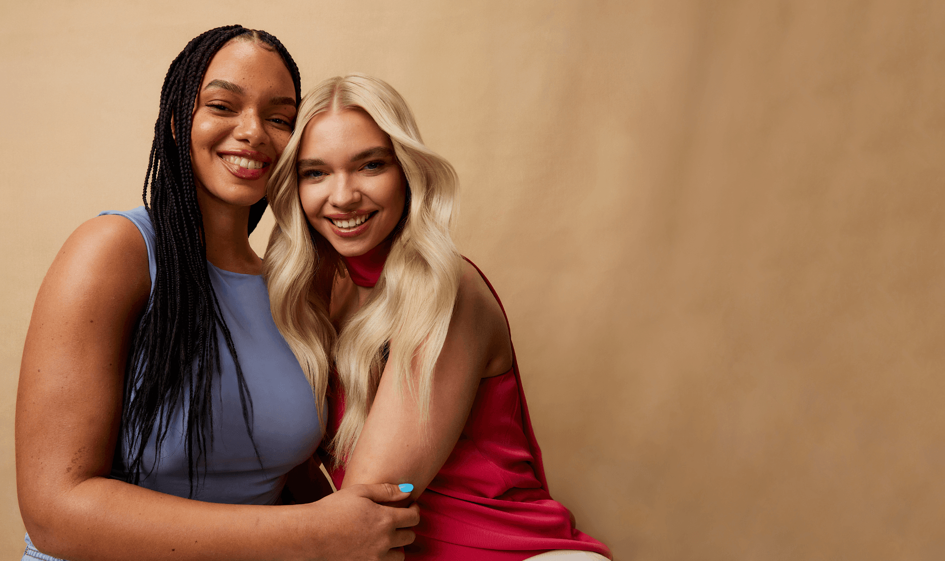  Two young women hugging each other, both have glowing skin and are smiling.