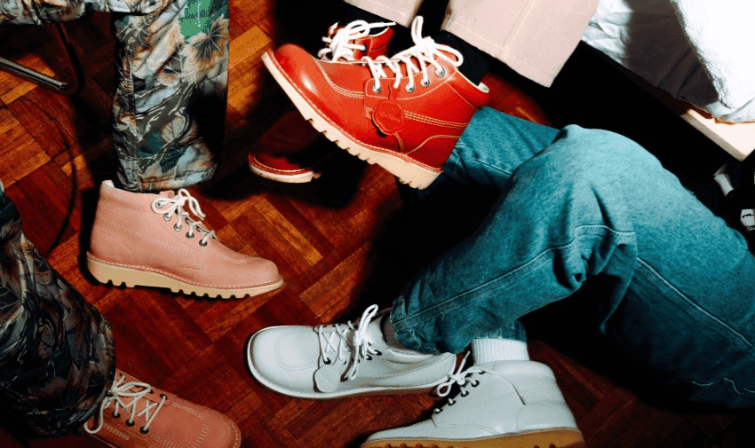 A group of people wearing a variety of colourful boots, including brown, red, and white, are seated together on a wooden floor. The image focuses on their footwear and lower legs, displaying various styles and patterns of pants along with the different boots