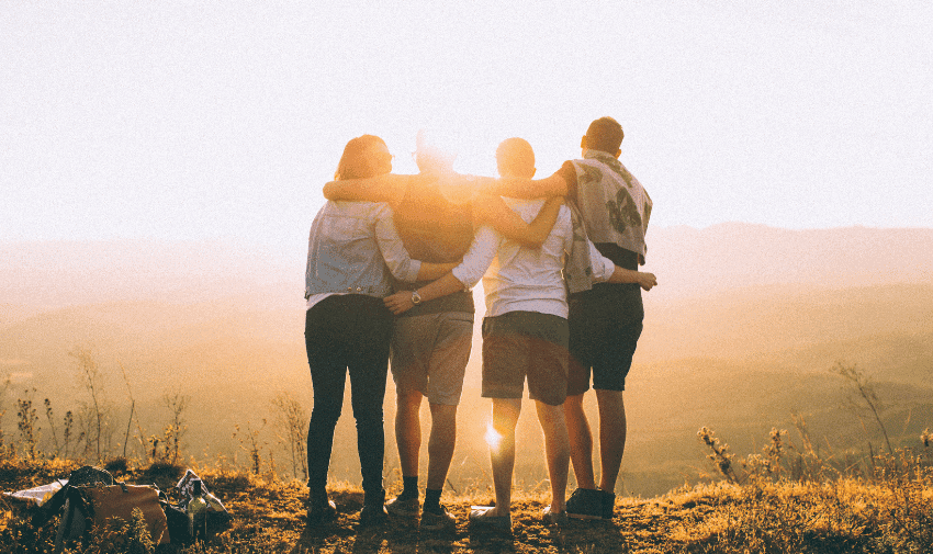 Four people are standing side by side with their arms around each other, facing a beautiful sunset on a hilltop. They are outdoors, with a scenic view of hills and valleys in the background. The sunlight casts a warm glow on the group.