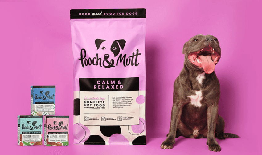 A happy brown dog with a white chest sits with its tongue out next to a large pink and purple bag of "Pooch & Mutt" Calm & Relaxed dog food. Three smaller containers of Pooch & Mutt dog food are stacked to the left. The background is pink.