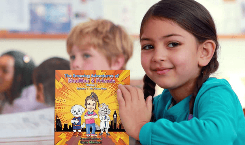 A young girl smiles while sitting at a desk, holding a colourful children's book titled "The Amazing Adventures of Moolmoo & Friends." The book cover shows illustrated animal characters in various poses against a vibrant backdrop. Other children are blurred in the background.