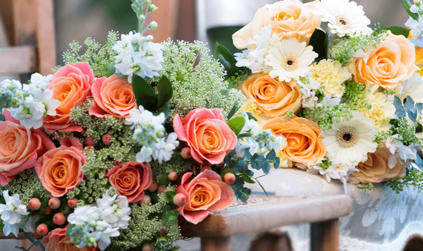 Two vibrant flower bouquets are displayed on wooden chairs. One has pink roses, white flowers, and green accents, while the other features yellow roses, white daisies, and greenery. Sunlight filters through, highlighting their colours and textures.