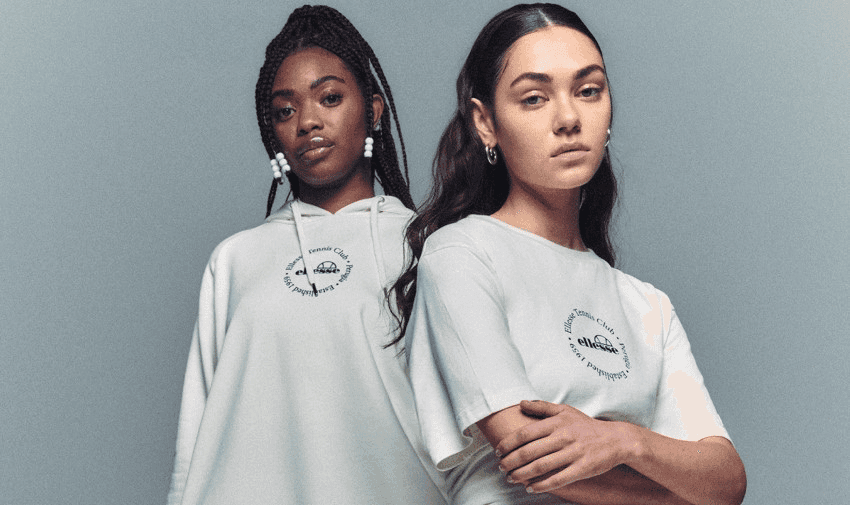 Two women stand side by side against a neutral background. The woman on the left has long braided hair and wears a light-coloured hoodie. The woman on the right has long straight hair and wears a light-coloured short-sleeve shirt. Both have a serious expression.