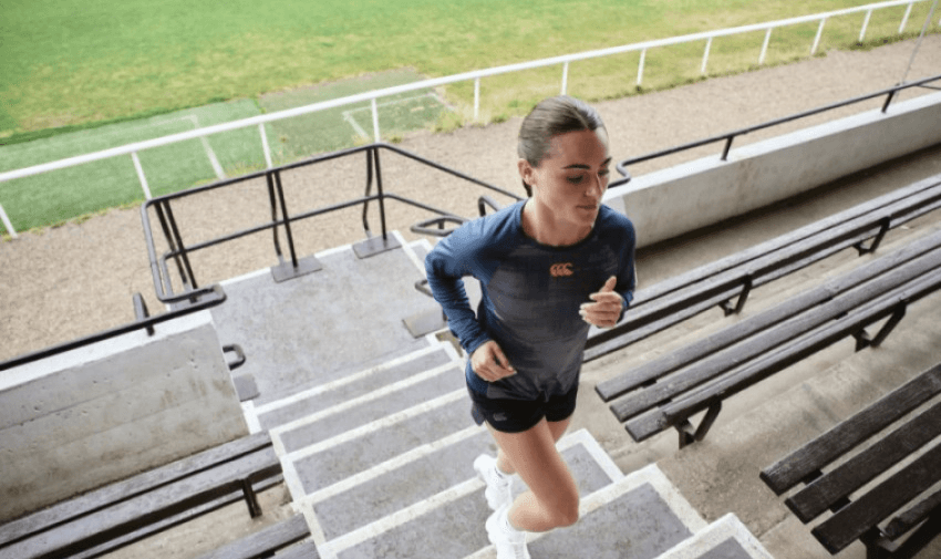 A person in athletic wear runs up stadium stairs, with green grass in the background. The setting is outdoors, with empty wooden benches on either side of the steps.