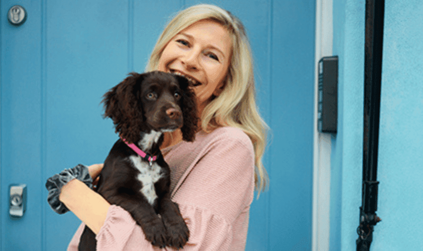 A person with long blond hair, wearing a pink long-sleeve top, smiles while holding a brown puppy with white markings and a pink collar. They are standing in front of a bright blue door.