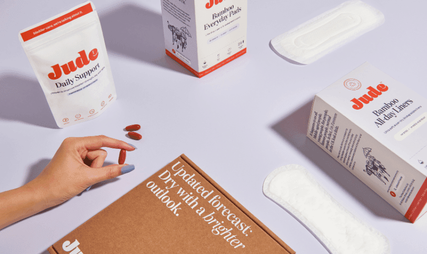 Top view of various feminine hygiene and health products arranged on a light purple surface. A hand is holding two pills next to a packet labeled "Jude Daily Support" and there are two boxes labeled "Jude Bamboo All-Day Liners" and sanitary pads placed around.