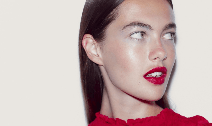 A person with long brown hair looks to the side against a light background. They are wearing a red top and have red lipstick. The image highlights their smooth skin and glossy lips.