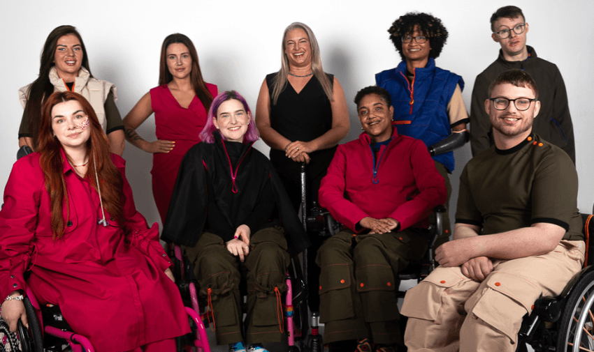 A diverse group of nine individuals, some in wheelchairs, are smiling and posing together. They are dressed in colourful and stylish clothing against a plain backdrop, conveying a sense of unity and inclusion.