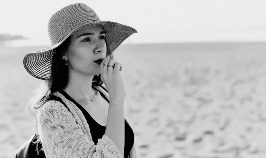 Black-and-white photo of a woman standing on a beach, wearing a large sunhat and light cardigan over a black top. She is looking into the distance with a thoughtful expression, with one hand near her mouth. The background is sandy and slightly blurred.