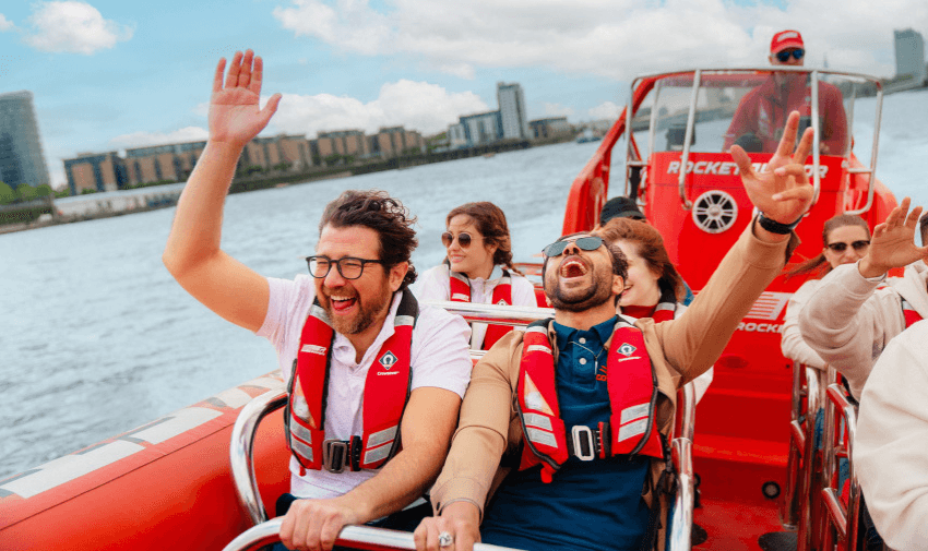 A group of people wearing life jackets are joyfully riding a speedboat on a river. Two men in the front are laughing and raising their arms in excitement. The background shows waterfront buildings under a partly cloudy sky.