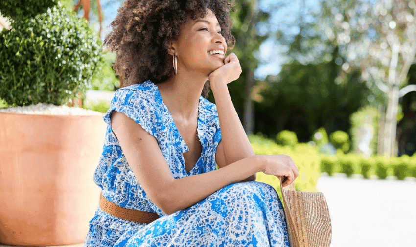 A woman with curly hair, wearing a blue and white floral dress, sits outdoors on a sunny day. She rests her chin on her hand and smiles while looking into the distance. She holds a straw hat in her lap, surrounded by greenery and sunlight.