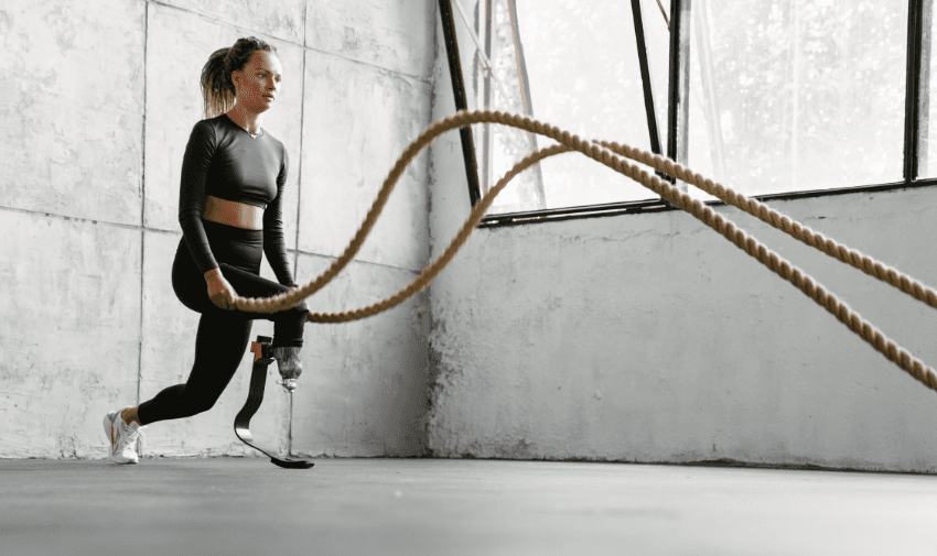 A woman with a prosthetic leg performing battle rope exercises in a minimalist gym, highlighting inclusivity and fitness. Discover disabled fitness discounts with Purpl