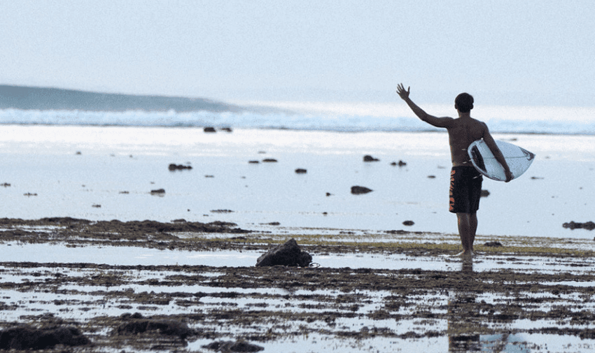 A man is walking towards the sea, he’s carrying a surfboard and has his left arm raised