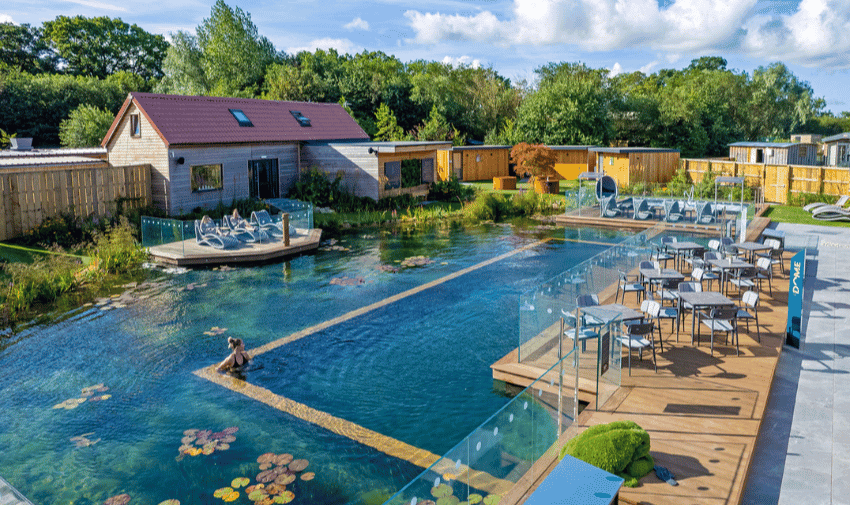 A serene outdoor spa setting features a swimming pool with a sunbather near its edge, surrounded by greenery and lily pads. Several seating areas with tables and chairs are lined up alongside the pool, with modern buildings and a wooden fence in the background.