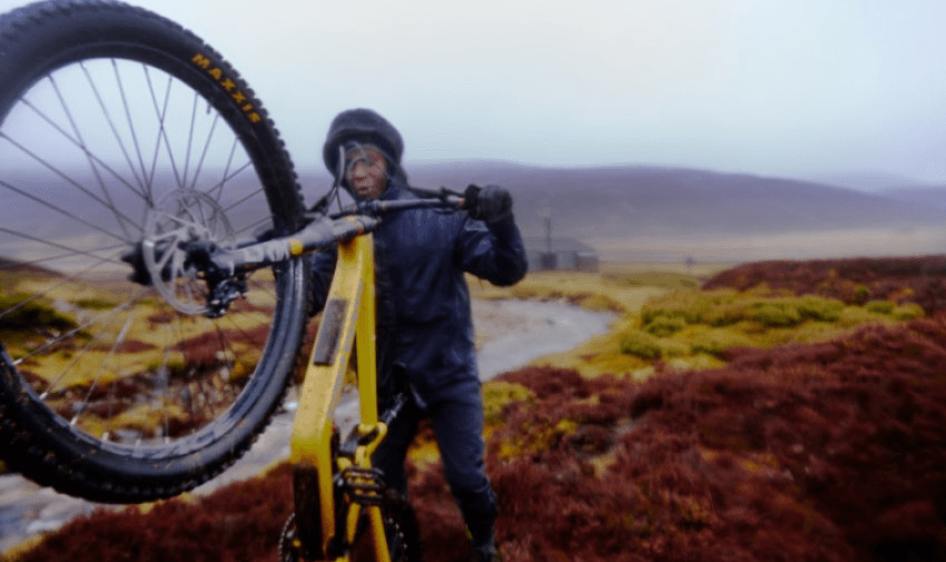 A person in wet-weather gear wheels a bicycle uphill in a misty, rugged landscape. They are framed by heather-covered ground and distant hills, with a grey sky overhead, suggesting a rainy or foggy day.