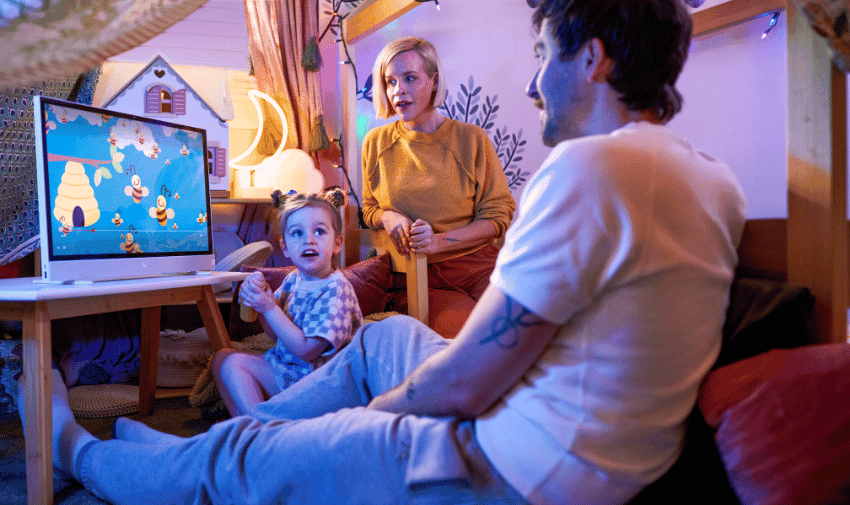 A family of three is gathered in a cosy, softly lit room. A young girl, sitting on the floor, is engaged with a computer screen showing a cartoon. An adult woman sits behind her, smiling, while an adult man is beside them, watching attentively.