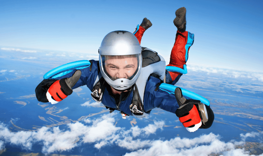 A person is skydiving, wearing a helmet, jumpsuit, and goggles, giving a thumbs-up with both hands. The background shows a clear, blue sky with scattered clouds and a distant view of land and water below, a perfect thrill for your next BuyAGift adventure!