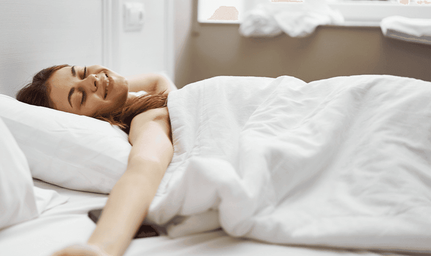 A person stretches and smiles while lying in bed under a white blanket. The room appears bright and airy with light coming in through a window.