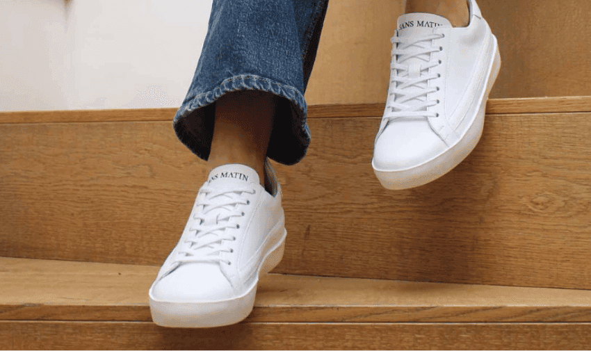 Person wearing white trainers with "Sans Matin" written on the tongue, descending wooden stairs. The person is dressed in blue jeans that slightly cover the tops of the sneakers. The background includes a wooden stairway