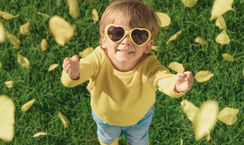 A young child with heart-shaped sunglasses, wearing a yellow sweater and blue pants, stands on green grass, smiling up at the camera. Yellow leaves are falling around them, suggesting a fun and playful autumn scene.