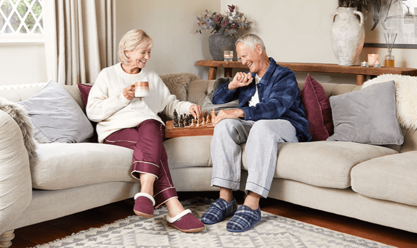 An elderly couple relaxing at home, wearing Cosyfeet slippers while enjoying a chess game, with a cosy and warm atmosphere. Promoting Purpl's disabled discounts for comfortable and adaptive footwear, perfect for all-day support in the UK.