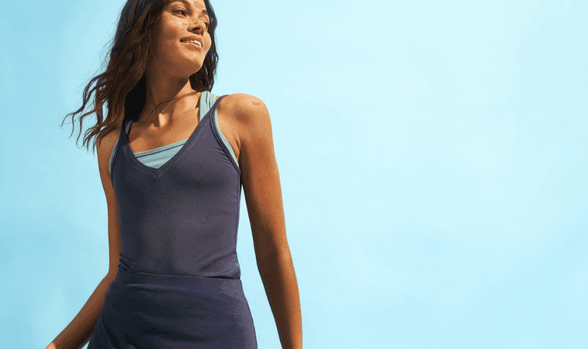 A woman with wavy brown hair is smiling and looking off to the side. She is wearing a dark blue tank top and a light blue sports bra underneath. The background is a clear blue sky.