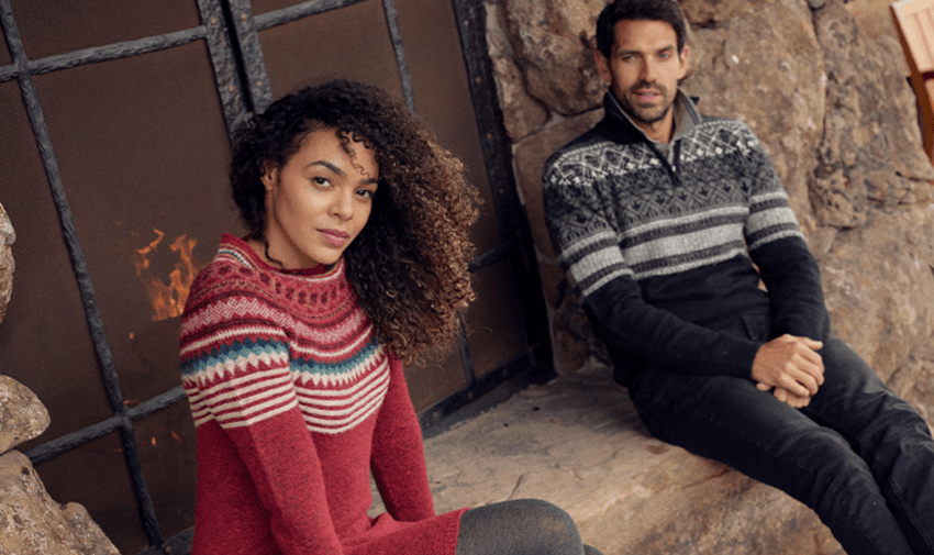 A woman in a red patterned sweater and a man in a dark patterned sweater are sitting by a stone fireplace. The woman is sitting on the floor, while the man is on a bench. The scene is cosy and warm.