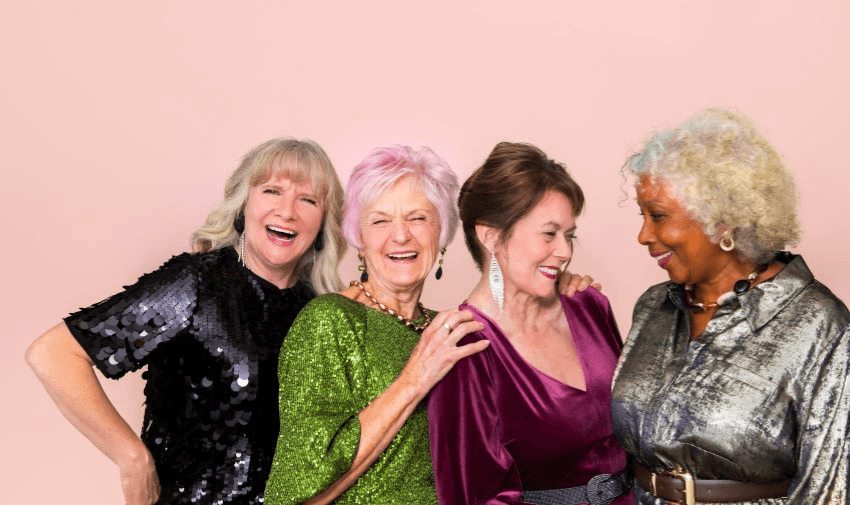 Four women smiling and laughing together against a pink background. They are dressed in shiny, elegant outfits, featuring sequins and velvet, exuding a joyful and glamorous vibe.