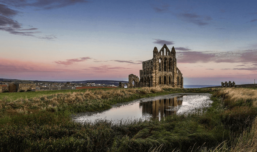 Ruins of a large stone abbey (Whitby Abbey) standing on a grassy hilltop at sunset, reflected in a small, still pond in the foreground. The sky is a blend of soft pink, purple, and blue hues, with distant views of a coastal town and the sea on the horizon. The scene is tranquil, evoking a sense of history and natural beauty.