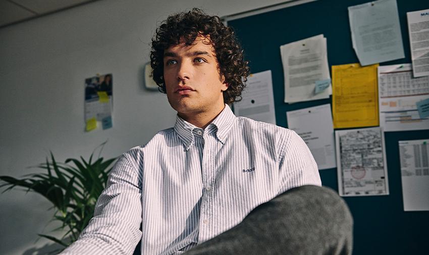 A person with curly hair sits in an office setting, wearing a striped shirt and looking pensively to the side. Papers and documents are pinned on the blue board behind them, and a potted plant is visible to the left.