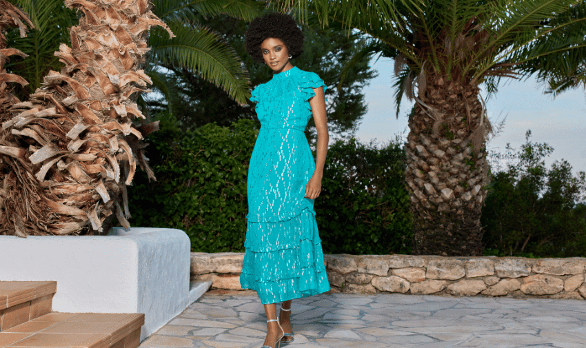 A woman with an afro hairstyle stands in a garden setting, wearing a vibrant blue, ruffled dress. She poses confidently on a stone pathway, surrounded by tall palm trees and greenery. A white structure is partially visible to the left.