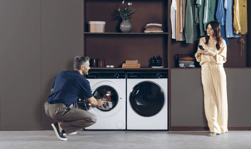 A man is kneeling and placing laundry into a Haier washing machine and tumble dryer, while a woman, standing to his right, looks at her phone.