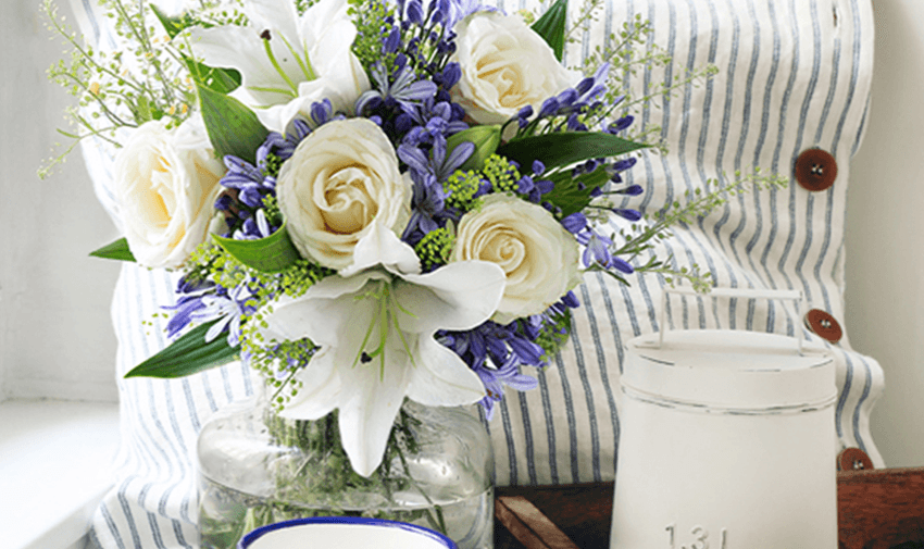 A bouquet of white roses, lilies, and purple flowers in a glass vase sits on a wooden surface. Nearby, a white candle is positioned next to a blue-rimmed dish. A striped pillow serves as the backdrop.