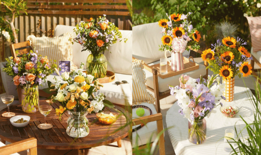 A sunny outdoor patio with wooden furniture, decorated with several vibrant floral arrangements. Bouquets of various flowers, including sunflowers and roses, are placed on the table and surrounding surfaces. Cushioned seating provides a comfortable and inviting atmosphere.