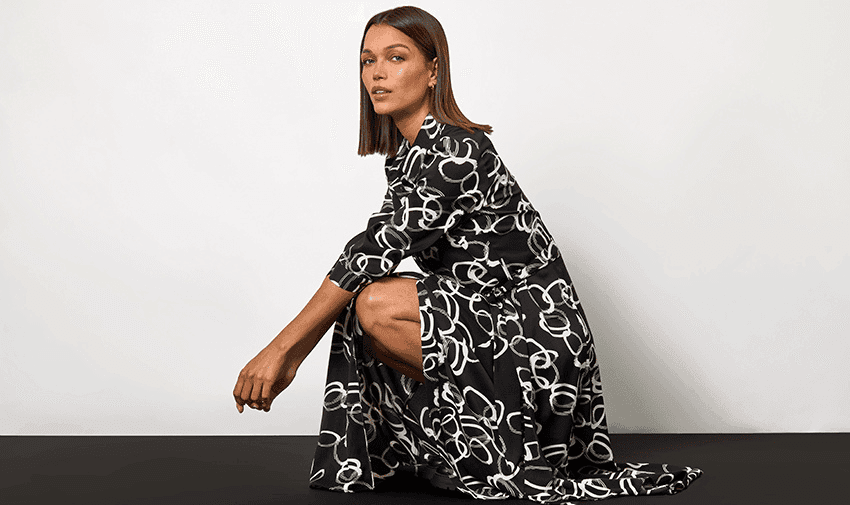 A person with straight, shoulder-length hair is wearing a black dress with white abstract patterns. They are crouching down on one knee with their hands resting on their other knee, set against a plain white background.