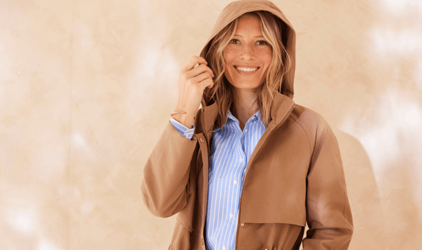 A smiling woman wearing a light brown hooded jacket over a blue and white striped shirt. She stands against a beige wall, gently holding her hood. Disabled fashion discounts UK, inclusive clothing deals for disabled people, accessible fashion offers.