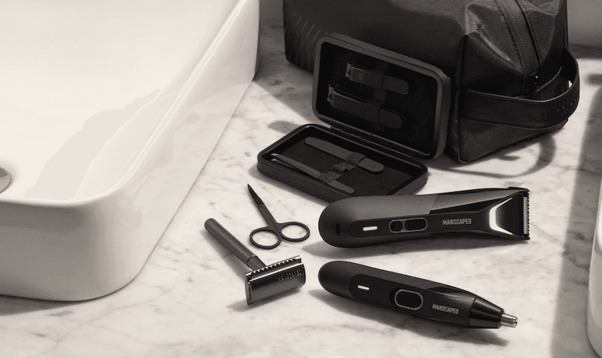 Manscaped grooming tools displayed on a marble countertop beside a white sink and a black toiletry bag. The tools include scissors, a razor, a trimmer, tweezers, and nail clippers, with some tools placed in an open black case