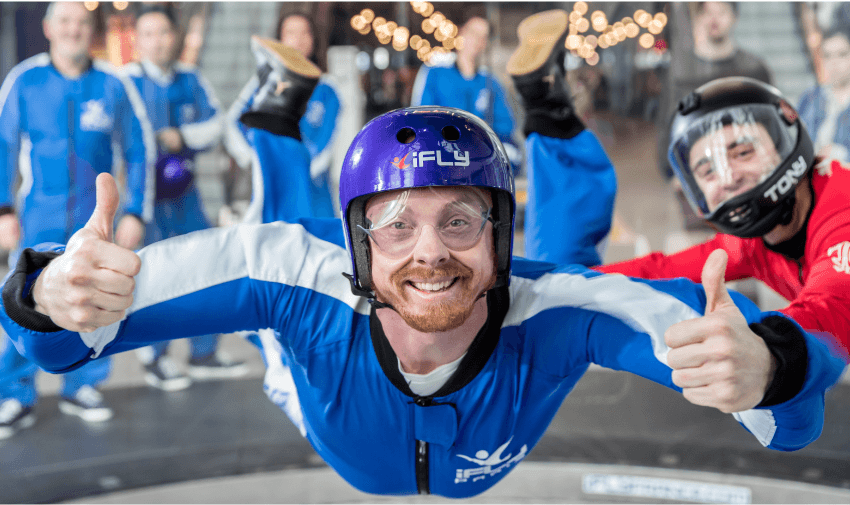 A smiling individual in a blue jumpsuit and helmet enjoying indoor skydiving at iFLY, surrounded by a supportive group. Purpl's disabled discounts make thrilling experiences like indoor skydiving accessible and inclusive for disabled individuals in the UK.