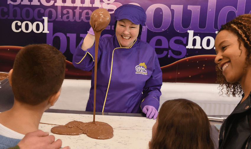 A Cadbury World chocolatier in a purple uniform skillfully pours melted chocolate onto a marble slab while smiling at a captivated audience. A fun and interactive chocolate-making experience for visitors of all ages. Cadbury World discounts for disabled people, UK attractions savings.