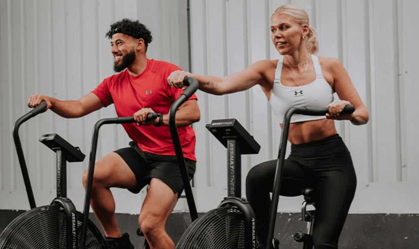 A man and a woman are working out on stationary bikes in a gym, wearing athletic Under Armour outfits, and smiling as they exercise. This image is for Sports Direct fitness gear discounts.