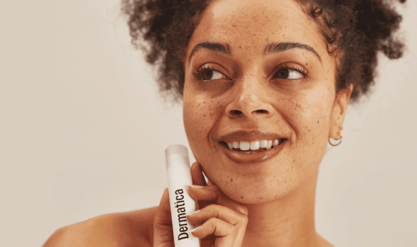 A woman with curly hair and freckles smiles while holding a tube of Dermatica skincare product. She has a natural look with minimal makeup and a warm skin tone. The background is plain and light-coloured.