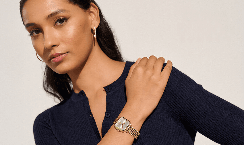 A person with long dark hair is wearing a navy ribbed top and a gold wristwatch. They have hoop earrings and are posing with one hand on their shoulder. The background is a plain light colour.