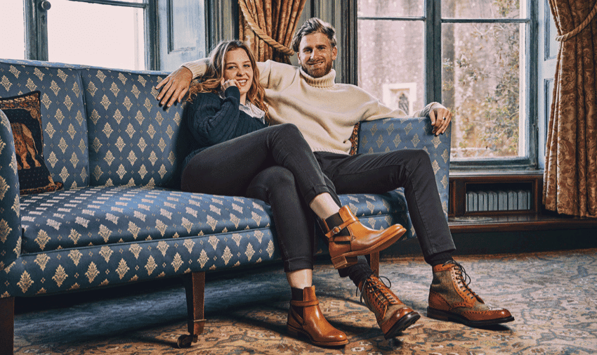 A smiling couple sits on a patterned blue couch. They are dressed in casual clothes and leather boots. The woman has her legs crossed on the couch.