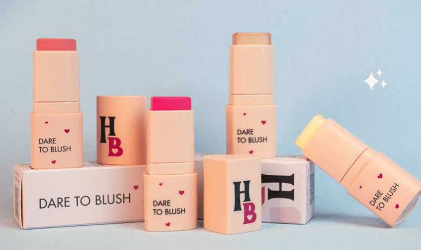 A variety of Human Beauty products, including blush sticks and hydrating lip glosses, arranged aesthetically against a pastel background, showcasing a playful and vibrant beauty brand ethos