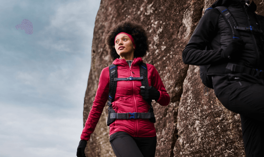 A person in a red jacket and gloves is rock climbing, looking upward with determination. Another climber in a dark jacket is partially visible. The sky is overcast, and the large rock face dominates the scene.